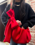 Person wearing a black sweater with a large red bow bag against a brick wall.
