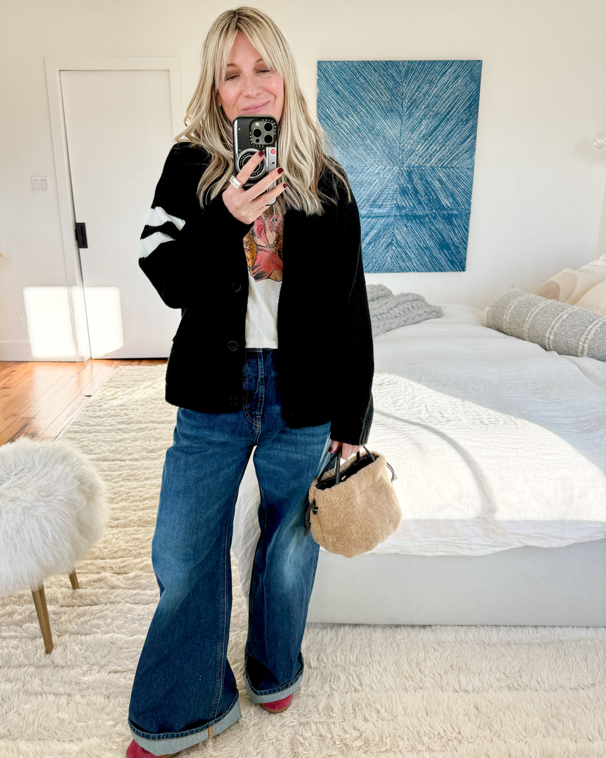 Selfie of a woman standing in a bedroom, wearing a black knit cardigan, graphic tee and jeans, holidng a beige fuzzy bag and wearing the East/West quartz statement cocktail ring in sterling silver.