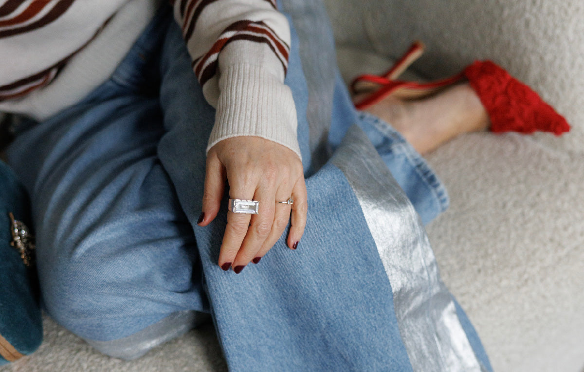 Person wearing a striped sweater and blue jeans with The East/West silver and clear quartz ring sitting on a couch with red shoes.