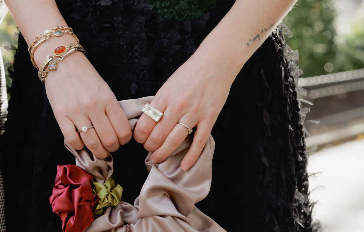 Person holding a beige clutch with floral details, wearing a black skirt, vintage watch band bracelets and a silver and quartz statement cocktail ring.