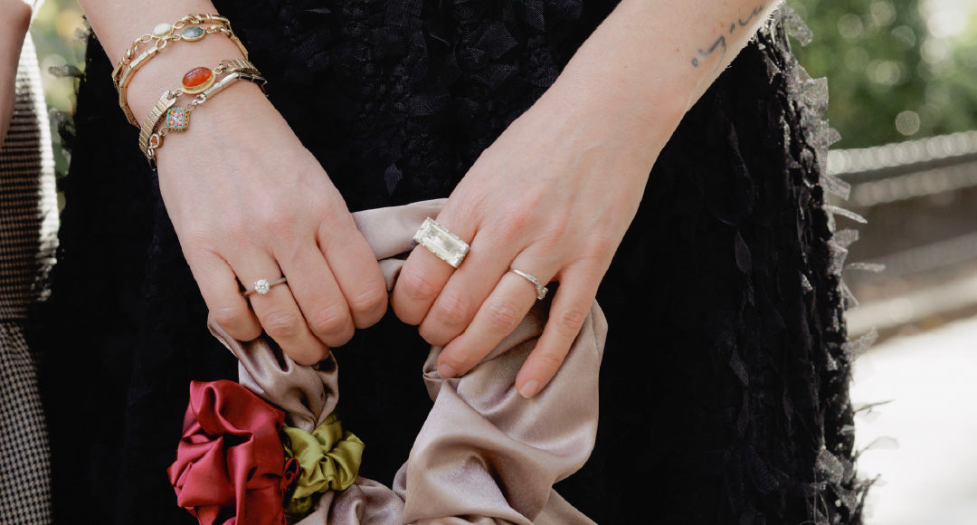 Person holding a beige clutch with floral details, wearing a black skirt, vintage watch band bracelets and a silver and quartz statement cocktail ring.