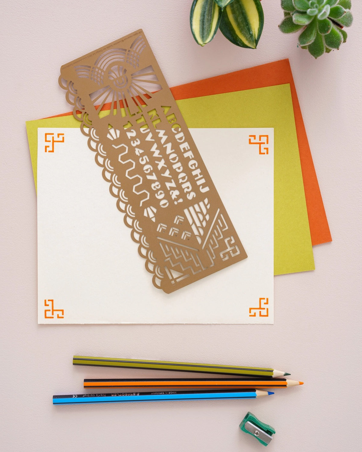 Brass stencil with a sunburst and alphabet laid on colorful paper beside pencils
