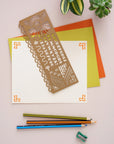 Brass stencil with a sunburst and alphabet laid on colorful paper beside pencils