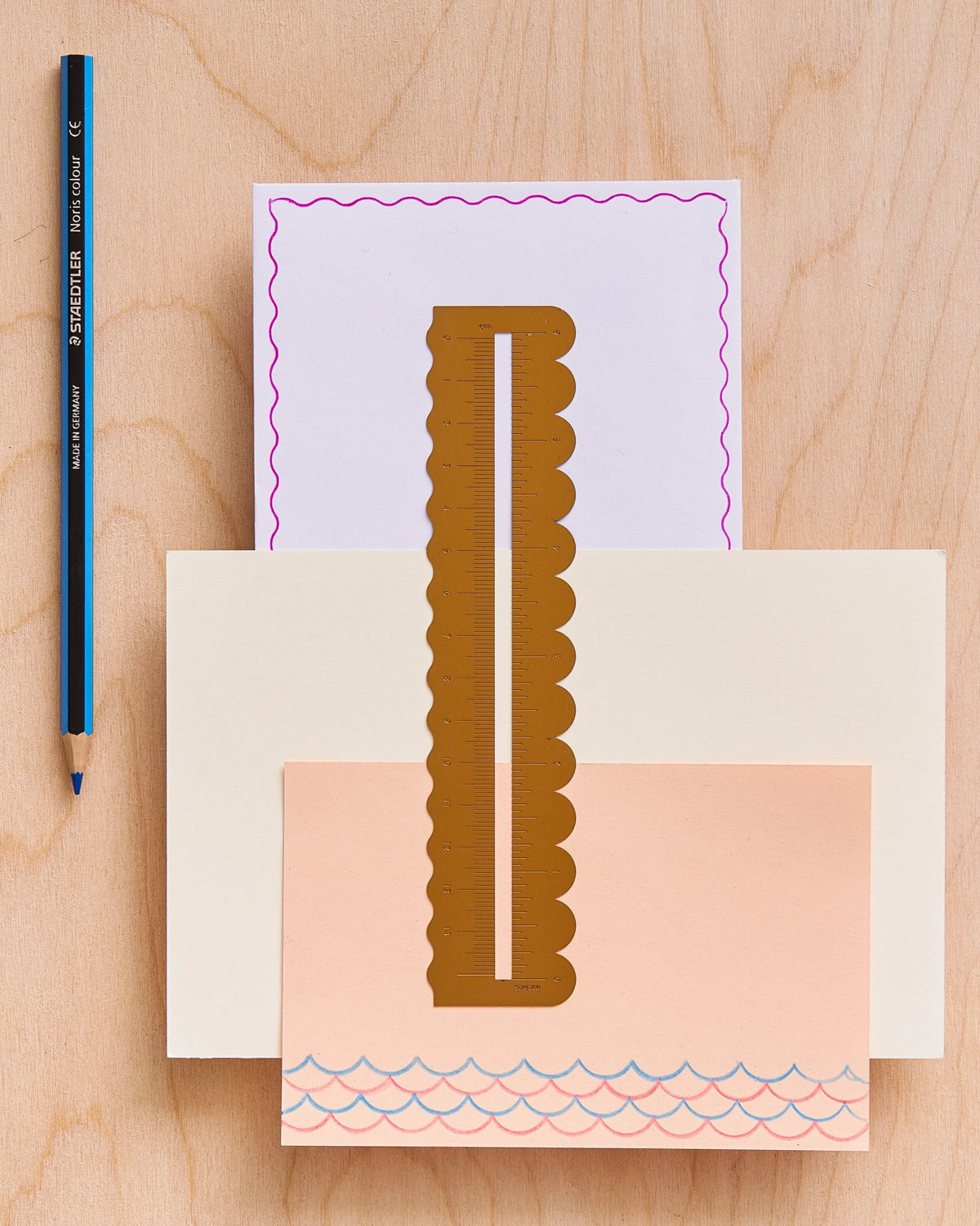 Brass scalloped ruler on layered pastel paper with a wavy drawn border and a blue pencil on a wood desk