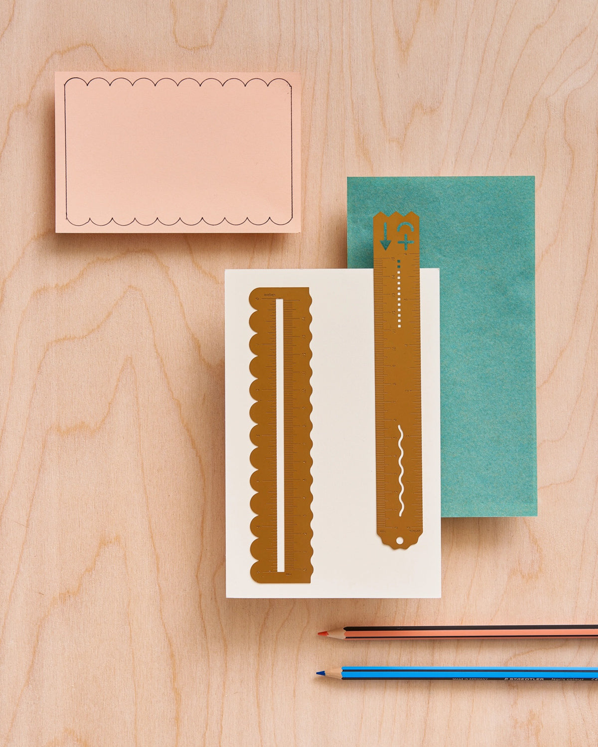 Brass wavy ruler on a cream card beside a scalloped peach note card, a teal envelope and a second brass ruler