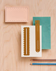 Brass wavy ruler on a cream card beside a scalloped peach note card, a teal envelope and a second brass ruler