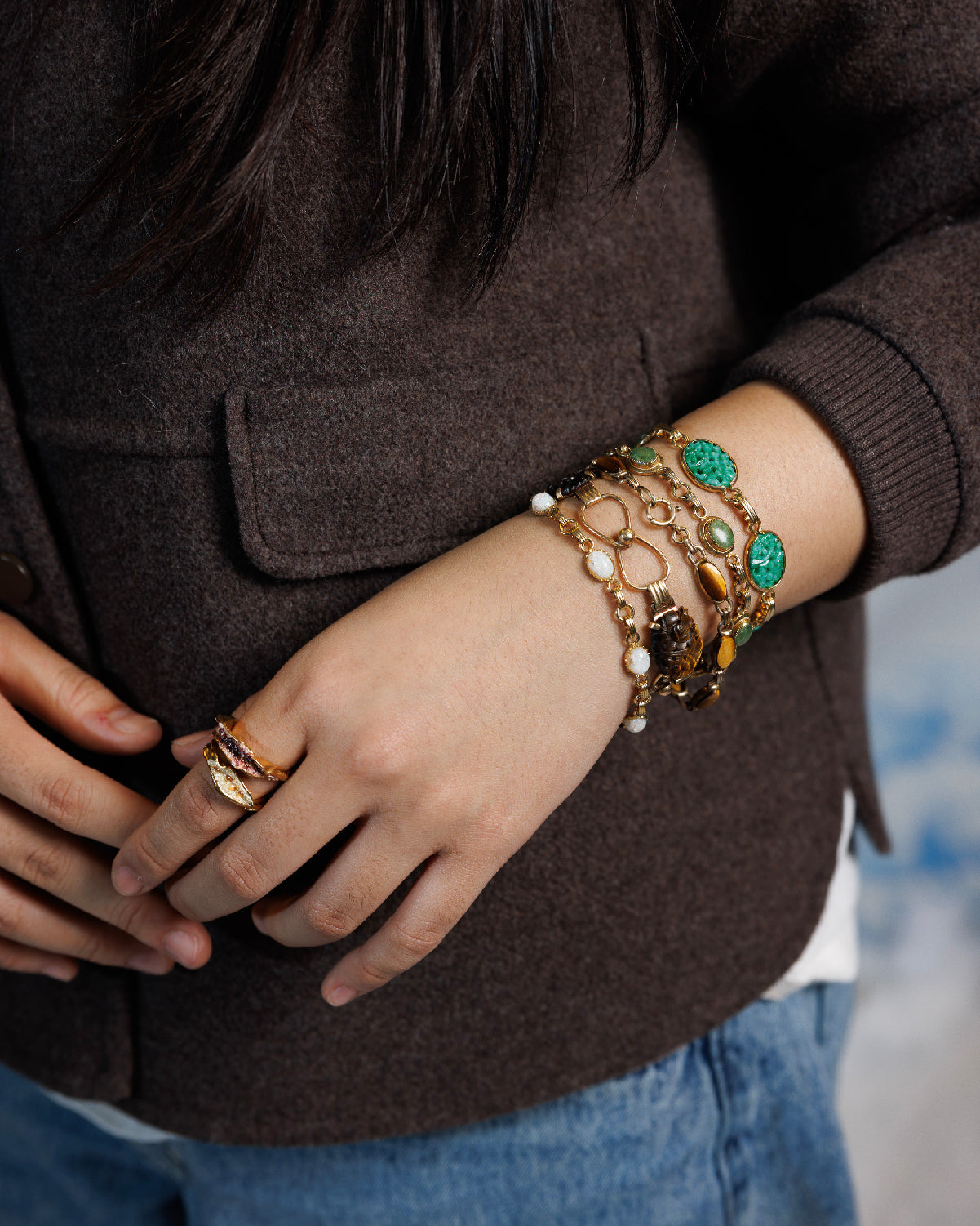 Close up of person's wrist and hand wearing 5 vintage gold-filled bracelets including tiger's eye, opal, carved peking glass, jade and two, gold rings with hand-painted glass centers.