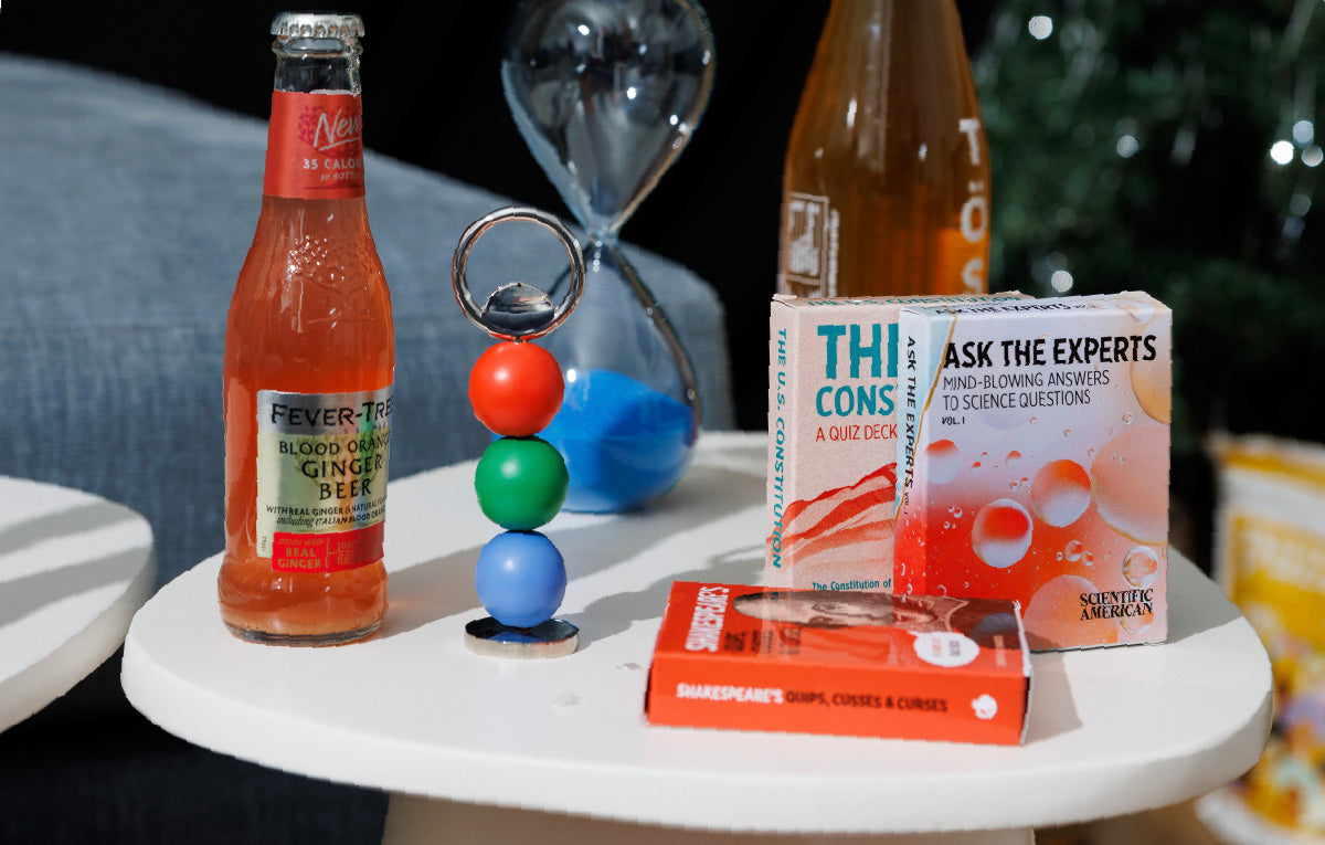 Ginger beer bottle on a table with Areaware Abaco bottle opener (red, green, blue), decks of cards and a blue hourglass.