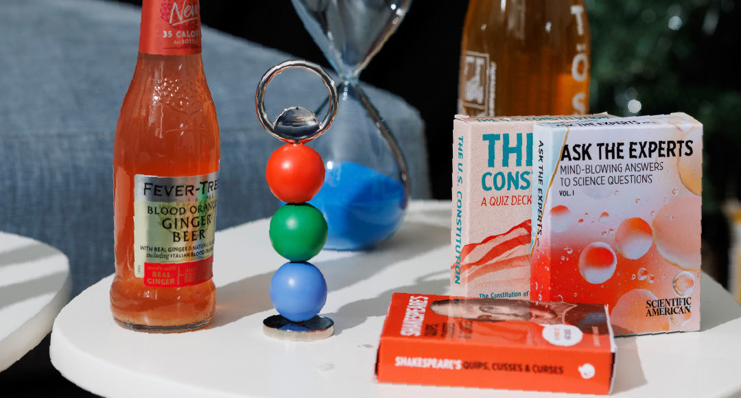 Ginger beer bottle on a table with Areaware Abaco bottle opener (red, green, blue), decks of cards and a blue hourglass.
