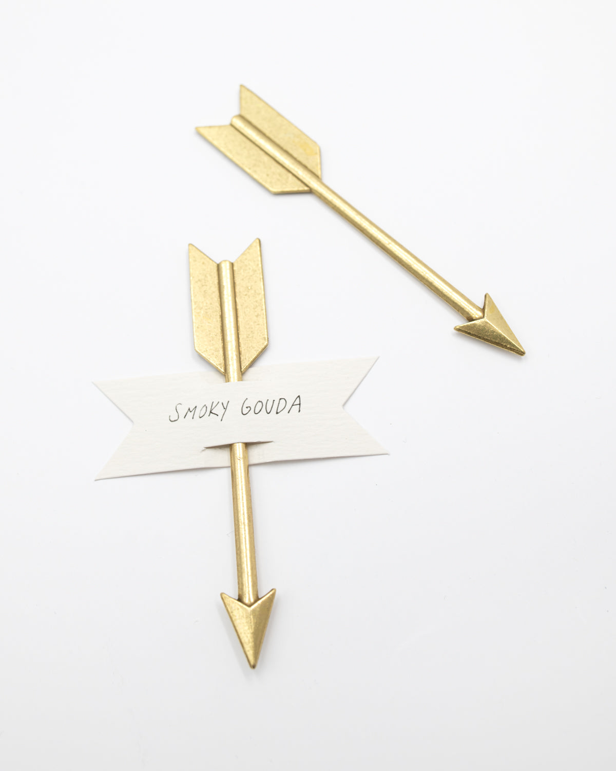 Two gold arrow cheese markers, one holding a paper banner that reads Smoky Gouda