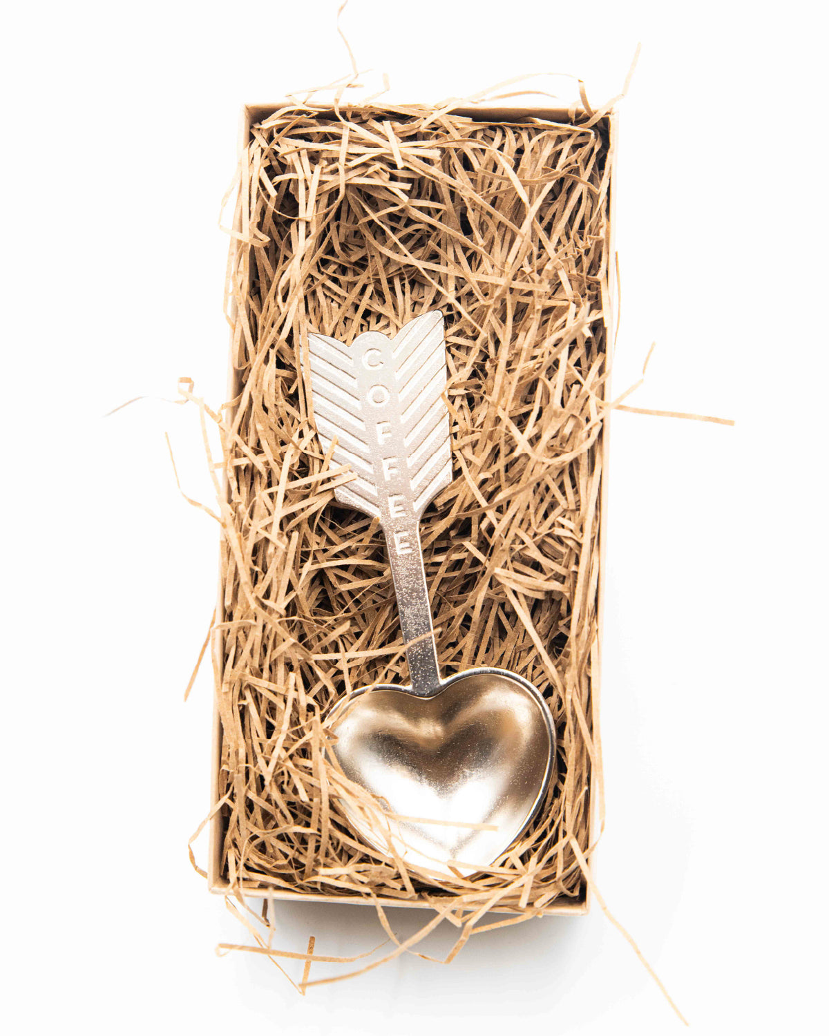 Metal coffee scoop with heart and arrow design in a box of straw on a white background.