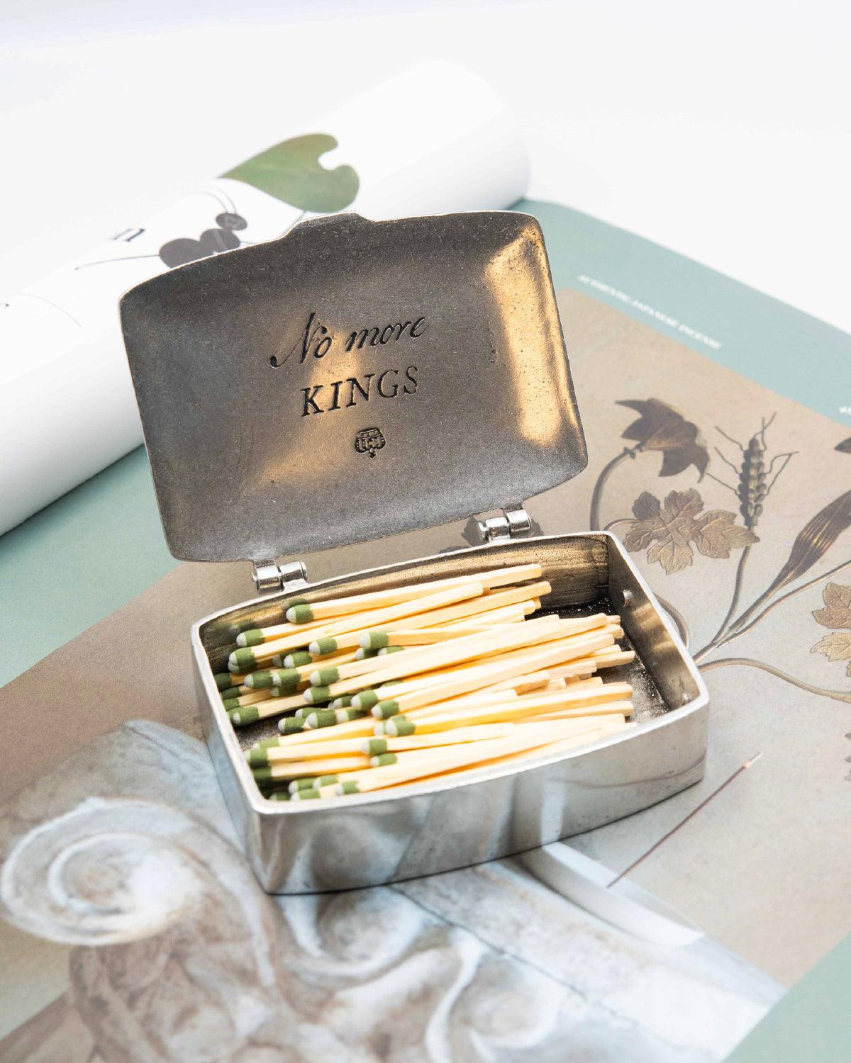 Beehive Handmade Spark a Revolution pewter matchbox open on botanical print magazine showing green-tipped strike-anywhere matches and "No More Kings" engraving inside lid