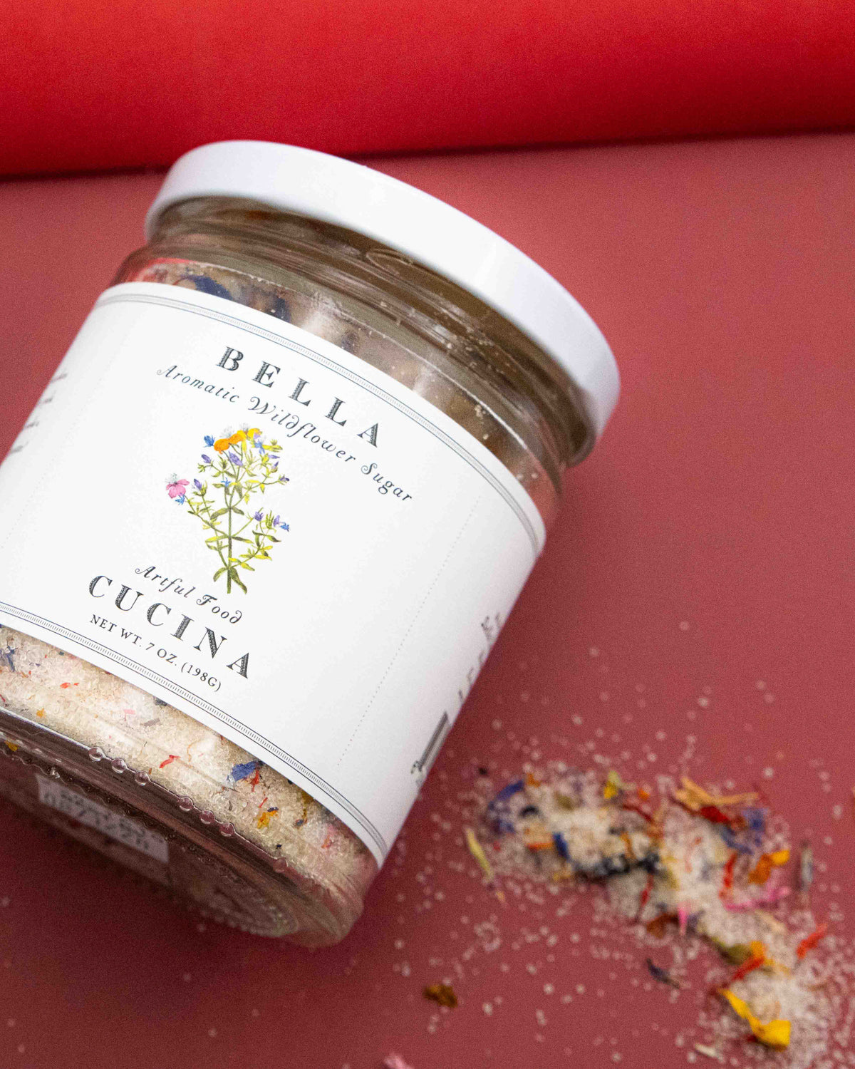 Jar of Bella Cucina Aromatic Wildflower Sugar with a botanical label with some sugar laying on the background.