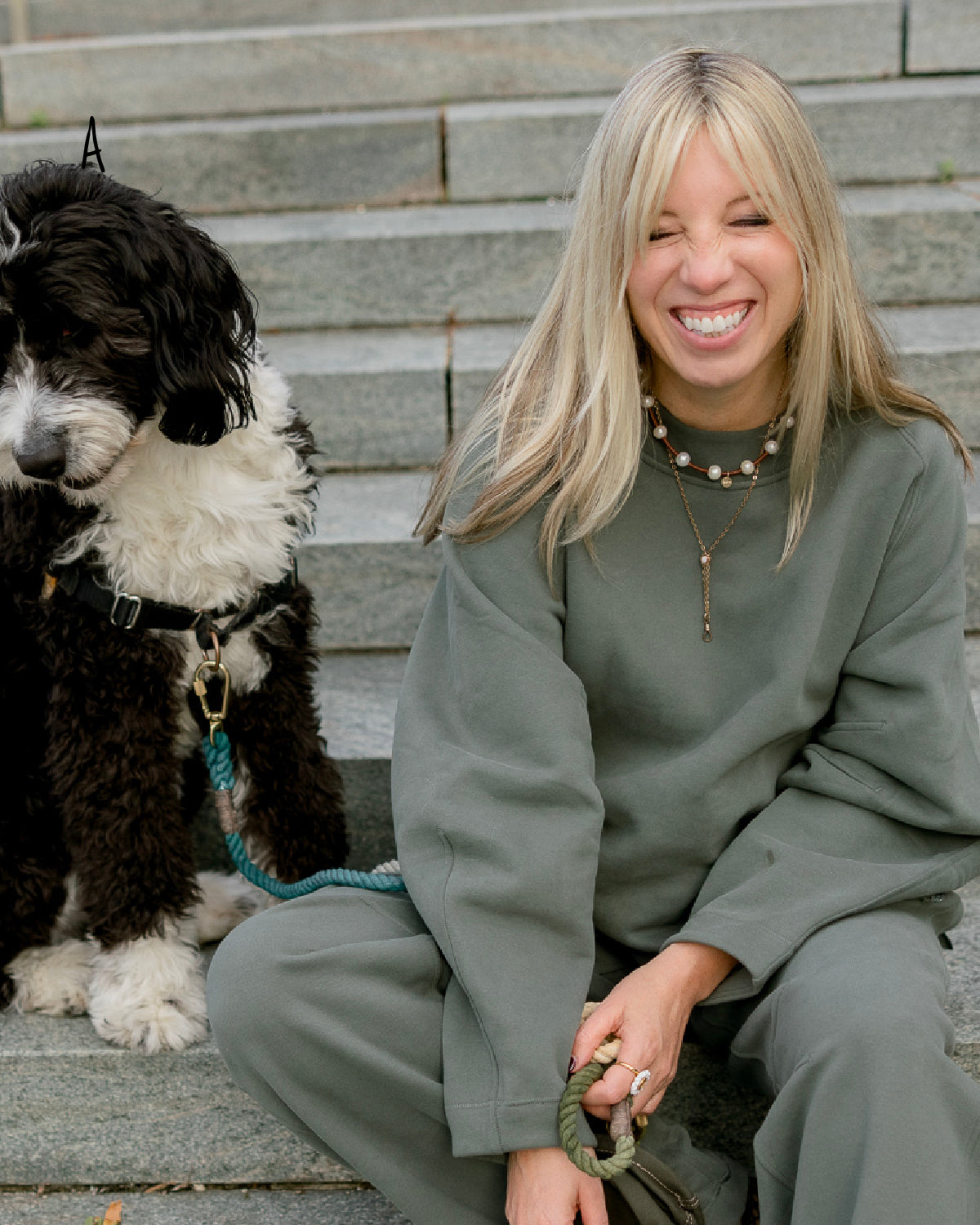 Woman sitting on steps wearing stacked pearl and leather necklace and vintage chatelaine necklace with a black and white dog on a rope leash