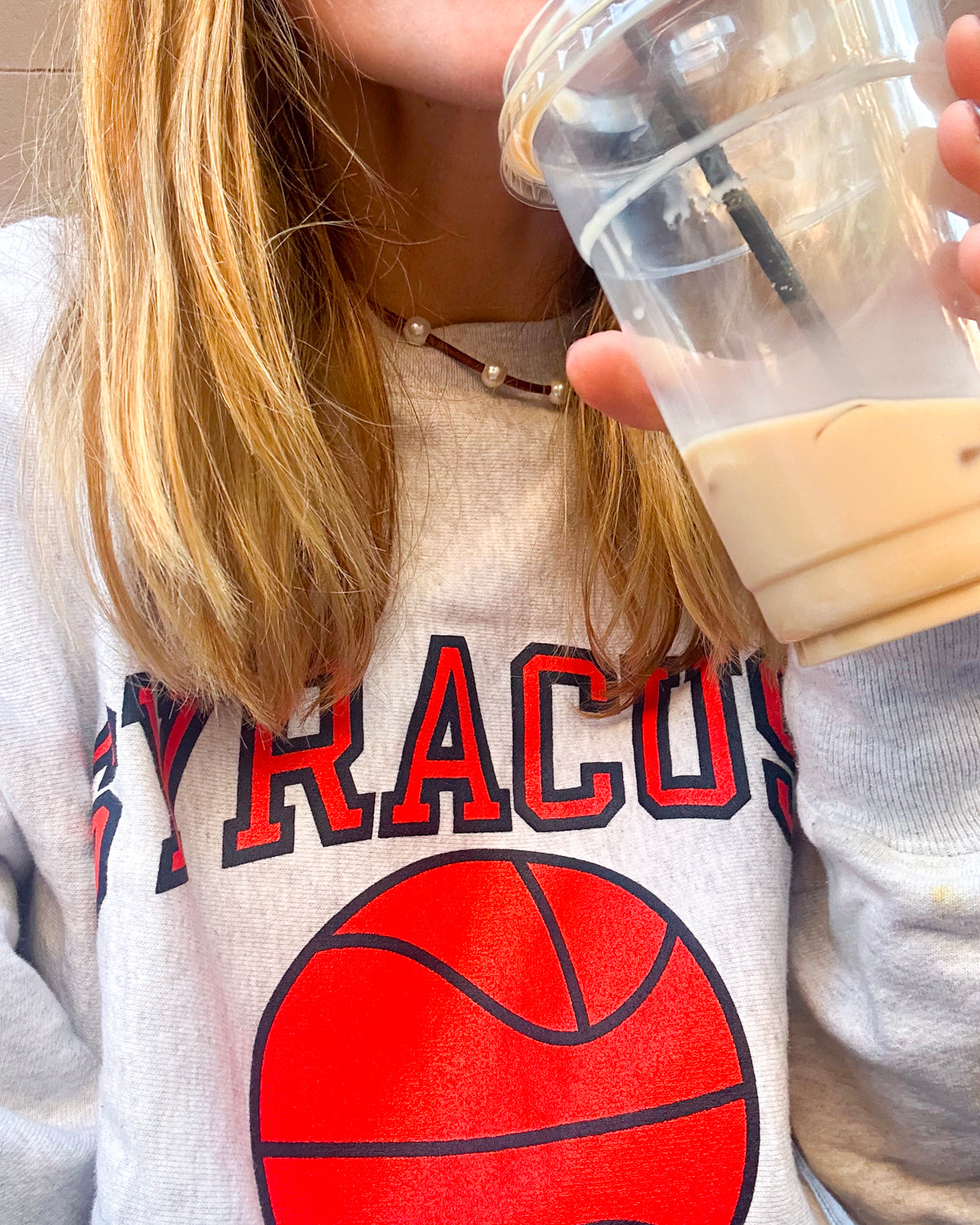 Close up of woman wearing pearl and leather cord necklace over a Syracuse basketball sweatshirt drinking iced coffee