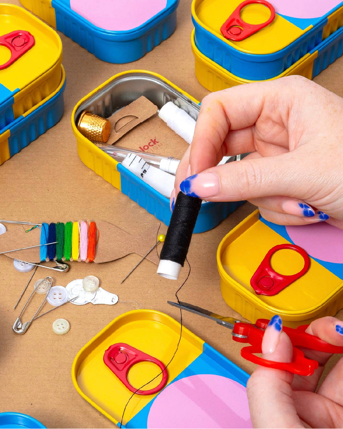Hands threading a needle over an open Sew Cute sewing kit tin with pins and colored thread