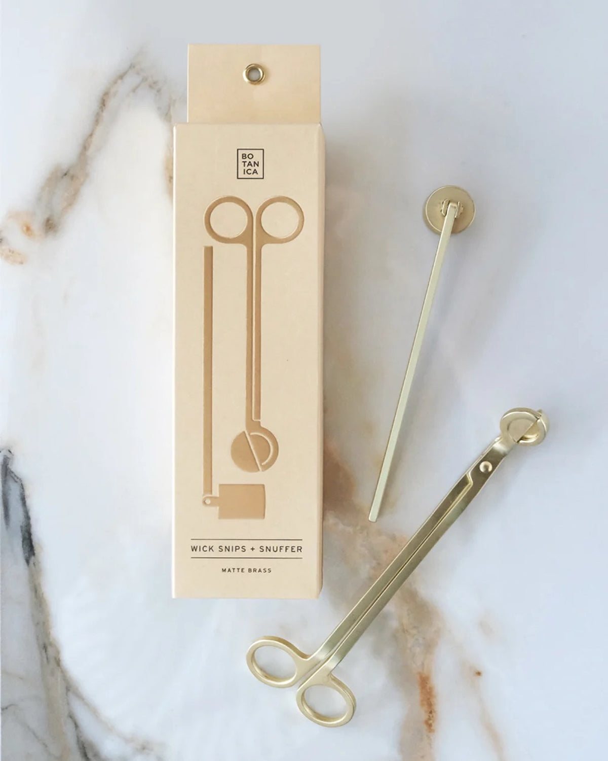 Matte brass candle wick snipper and snuffer next to their cream Botanica box