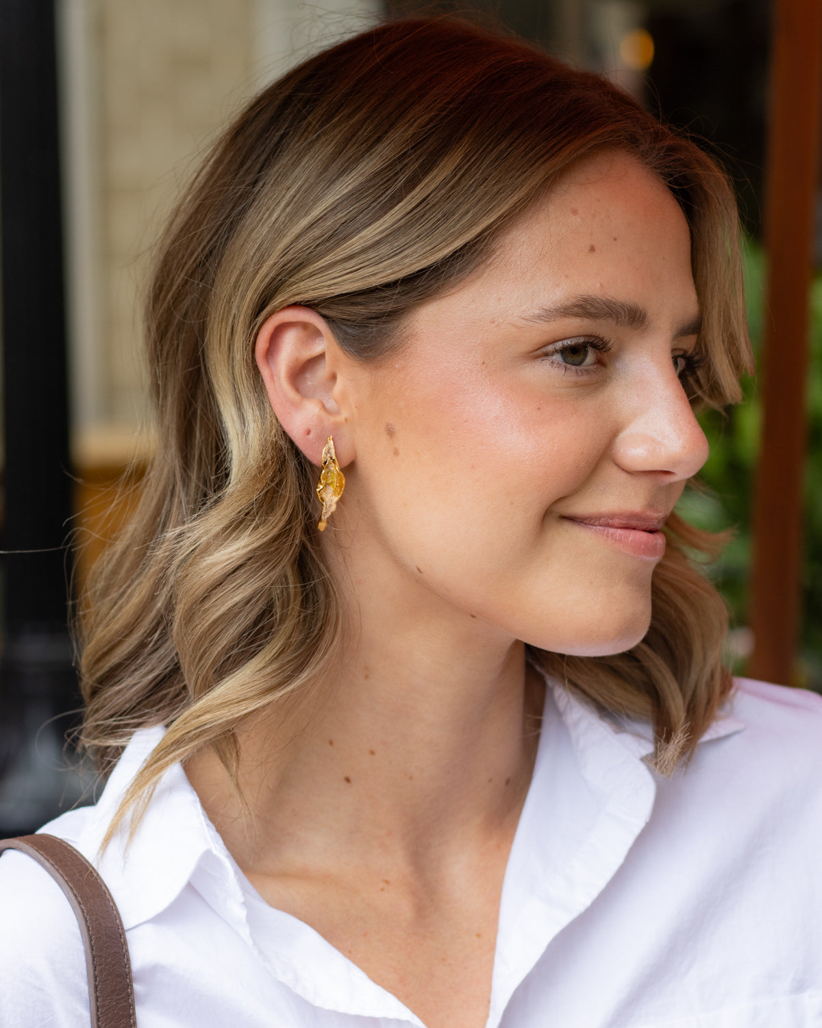 A woman with blonde-highlighted shoulder-length hair wearing a white collared shirt and a gold leaf-shaped dangle earring, shown in a close-up profile view.