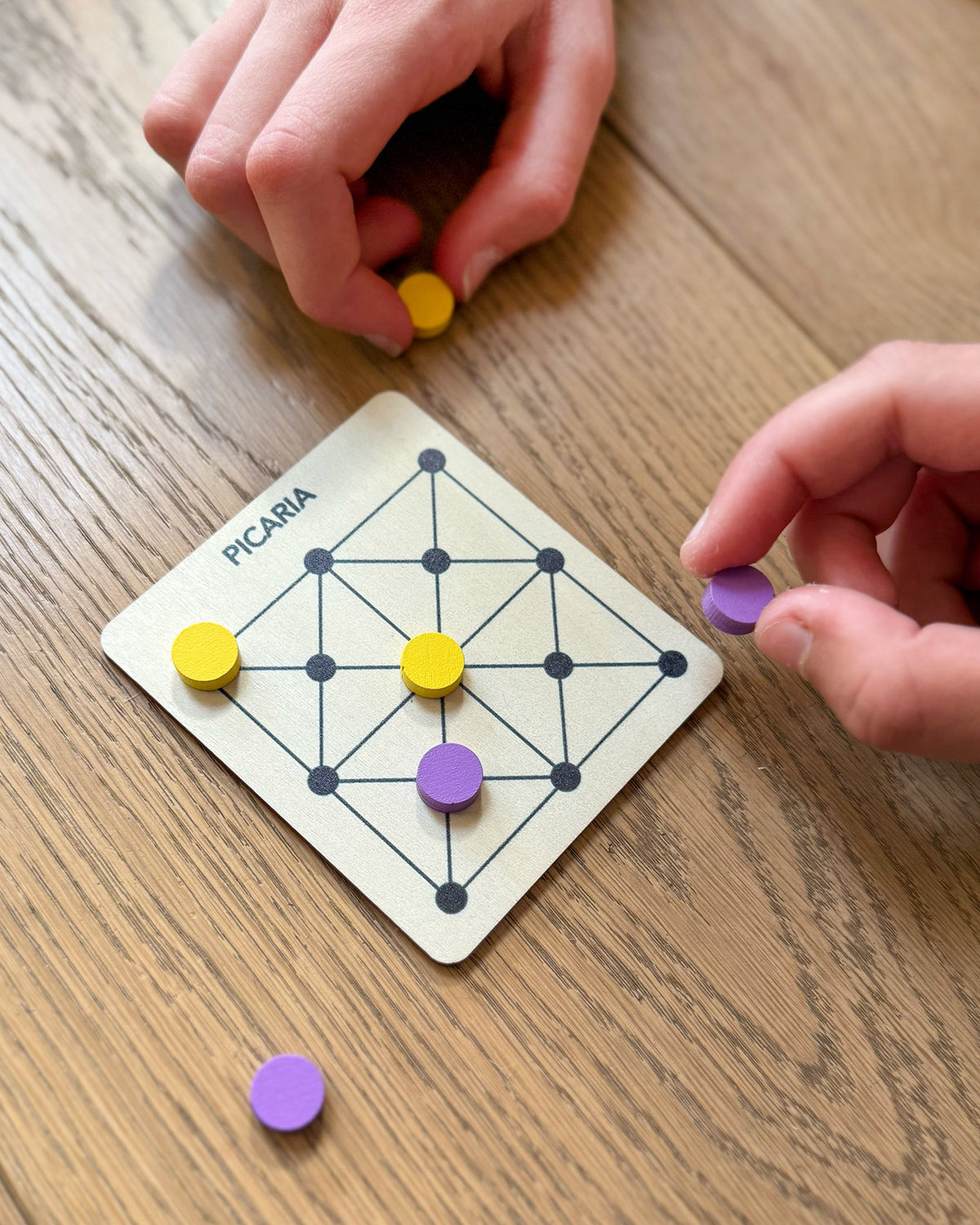 Hands playing Picaria with yellow and purple tokens on a square wooden board