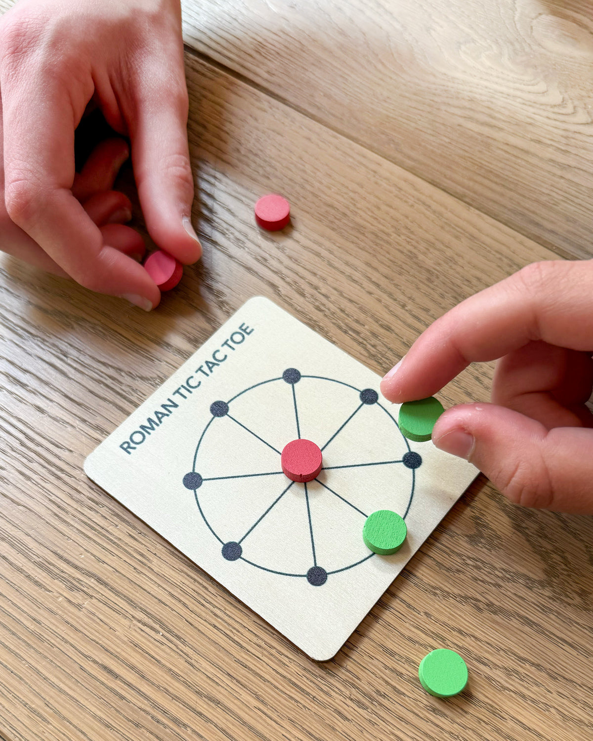 Hands playing Roman Tic Tac Toe with red and green tokens on a wooden board