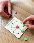 Hands playing Roman Tic Tac Toe with red and green tokens on a wooden board