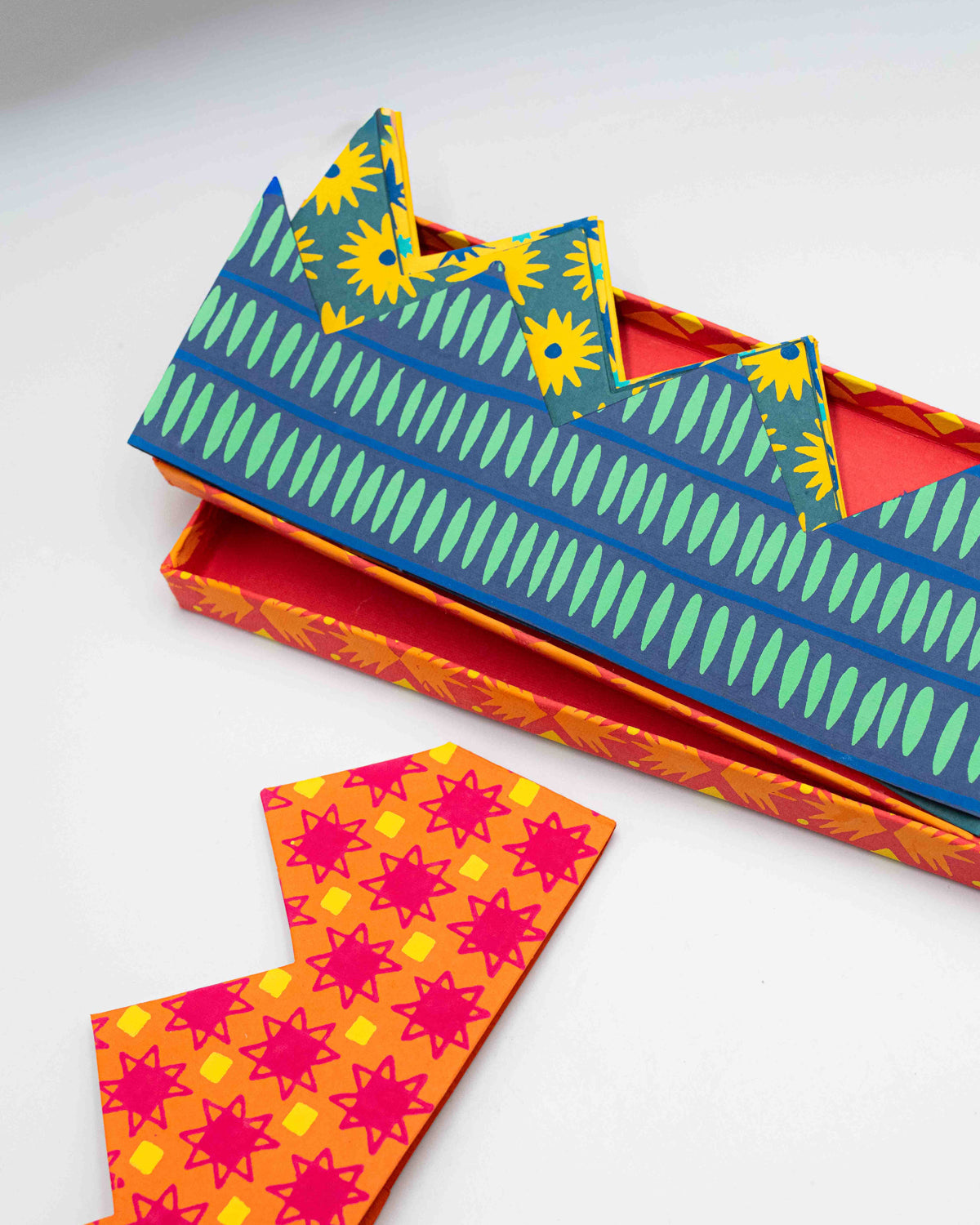 Blue and green striped paper crown on its orange gift box beside a red print crown