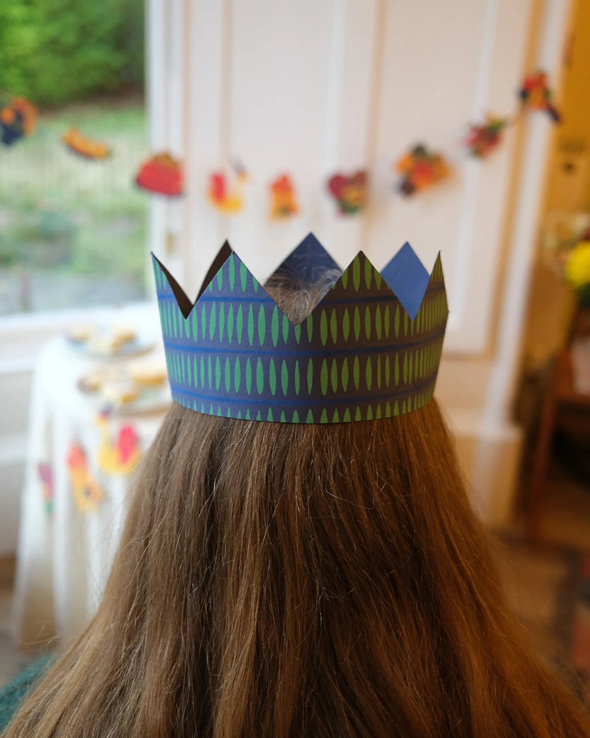 Child seen from behind wearing a navy and green paper crown in a festive room