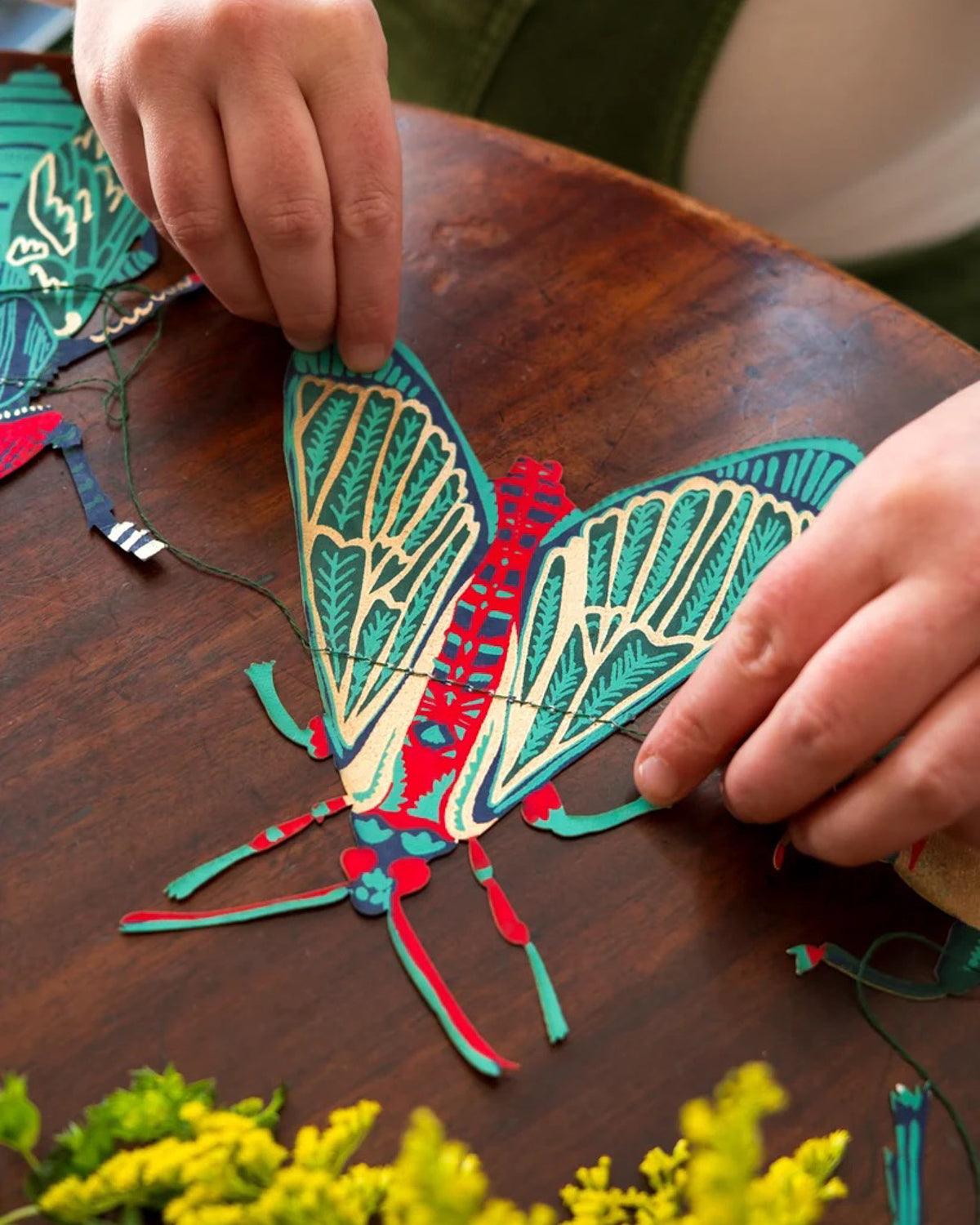 Hands threading a hand-printed paper moth in teal, red and gold onto the garland string