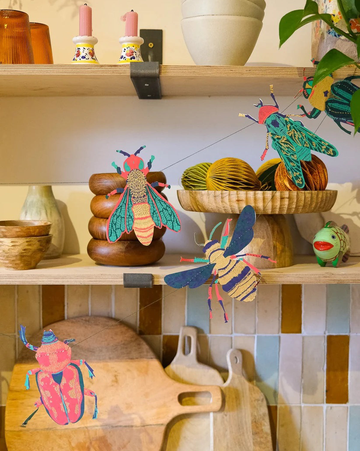 Paper bees and beetles strung across open kitchen shelves with wooden boards and pottery