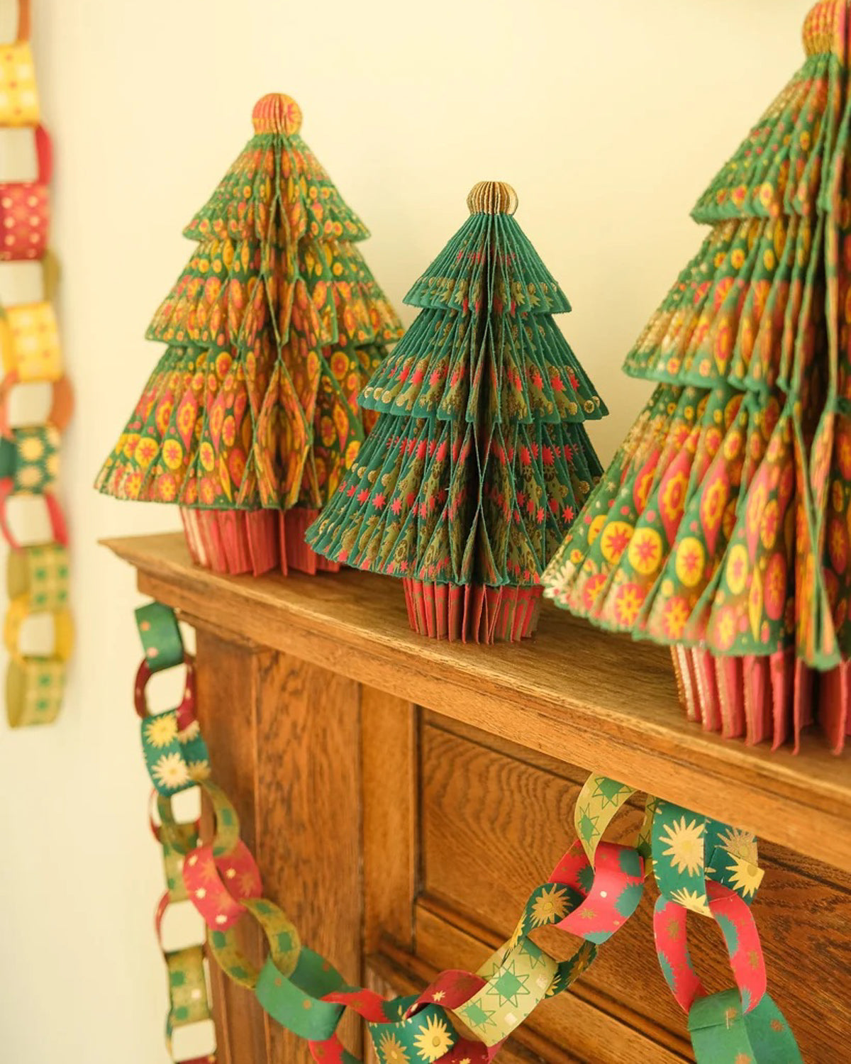 Patterned paper chains draped along a wooden mantel beside green paper Christmas trees
