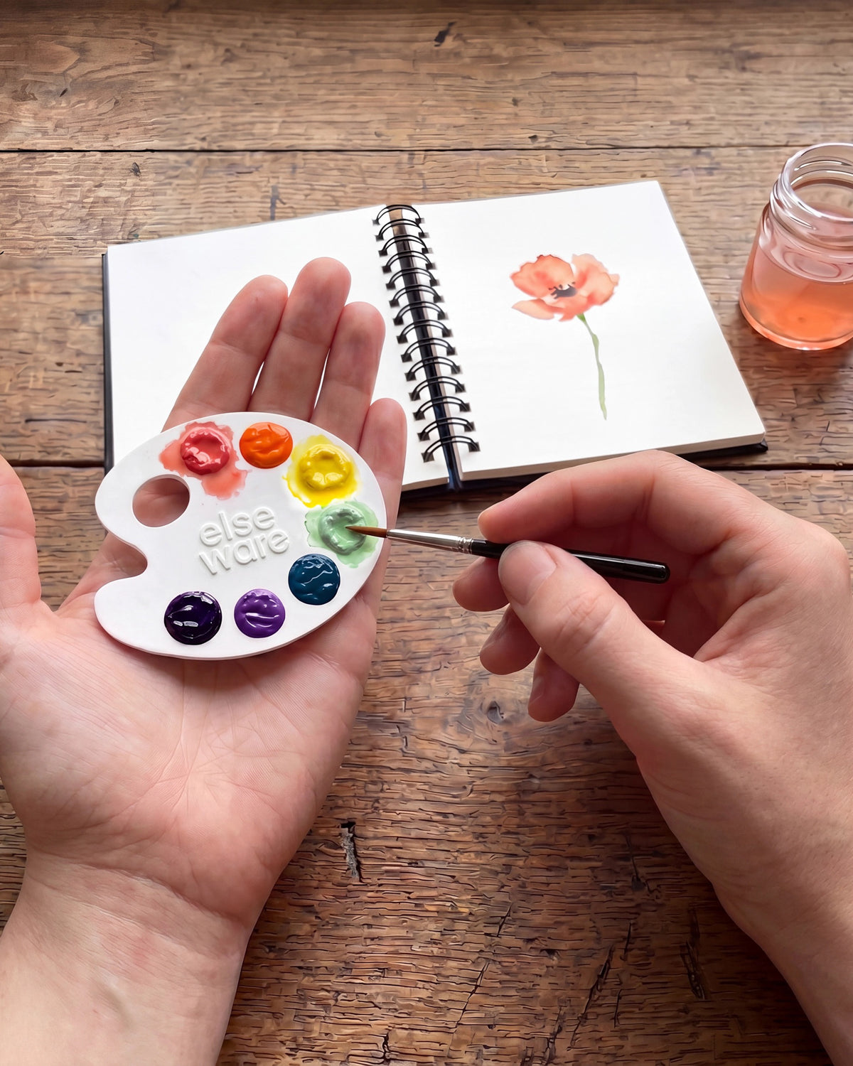 Hand holding a mini paper watercolor palette while painting a poppy in a sketchbook