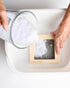 Hands pouring paper pulp into a wood mold set in a tub of water
