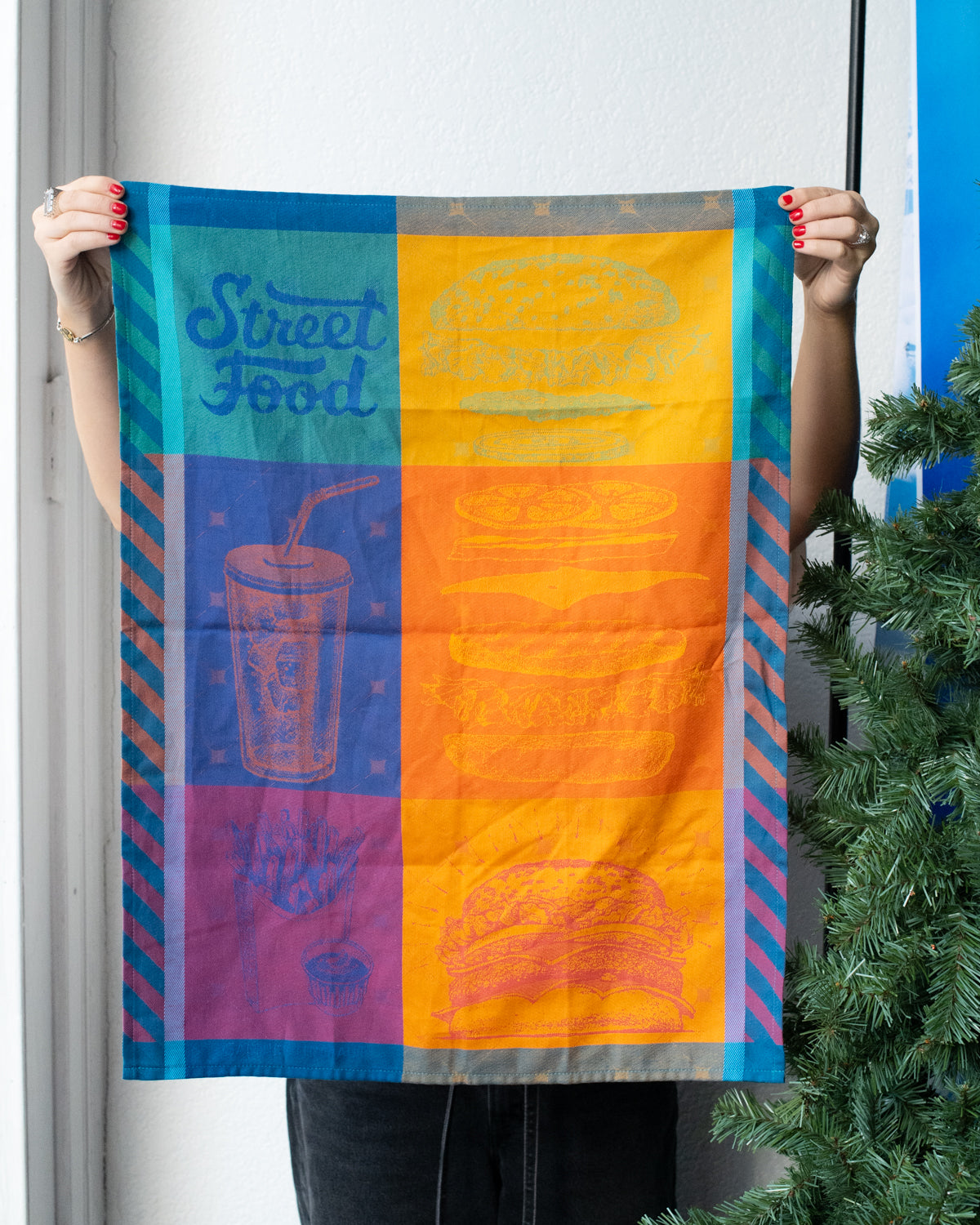 Person holding up the Street Food jacquard kitchen towel with burger, soda and fries graphics in teal and orange