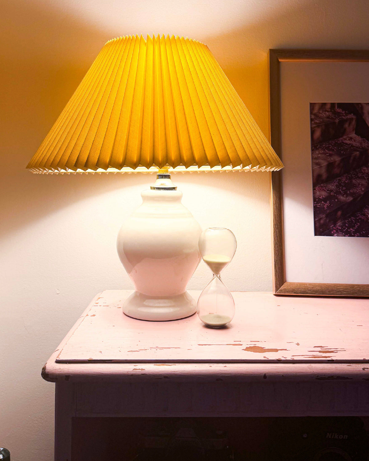 Small glass hourglass on a pink nightstand next to a lamp with a yellow pleated shade