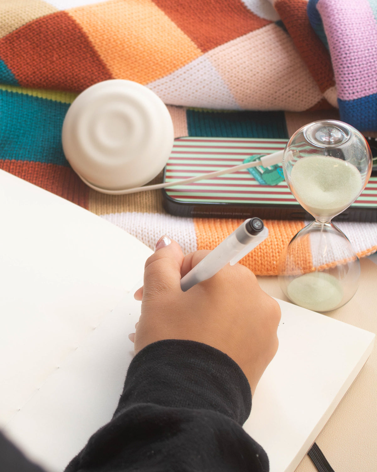 Hand writing in a notebook beside a glass hourglass and a striped pencil case