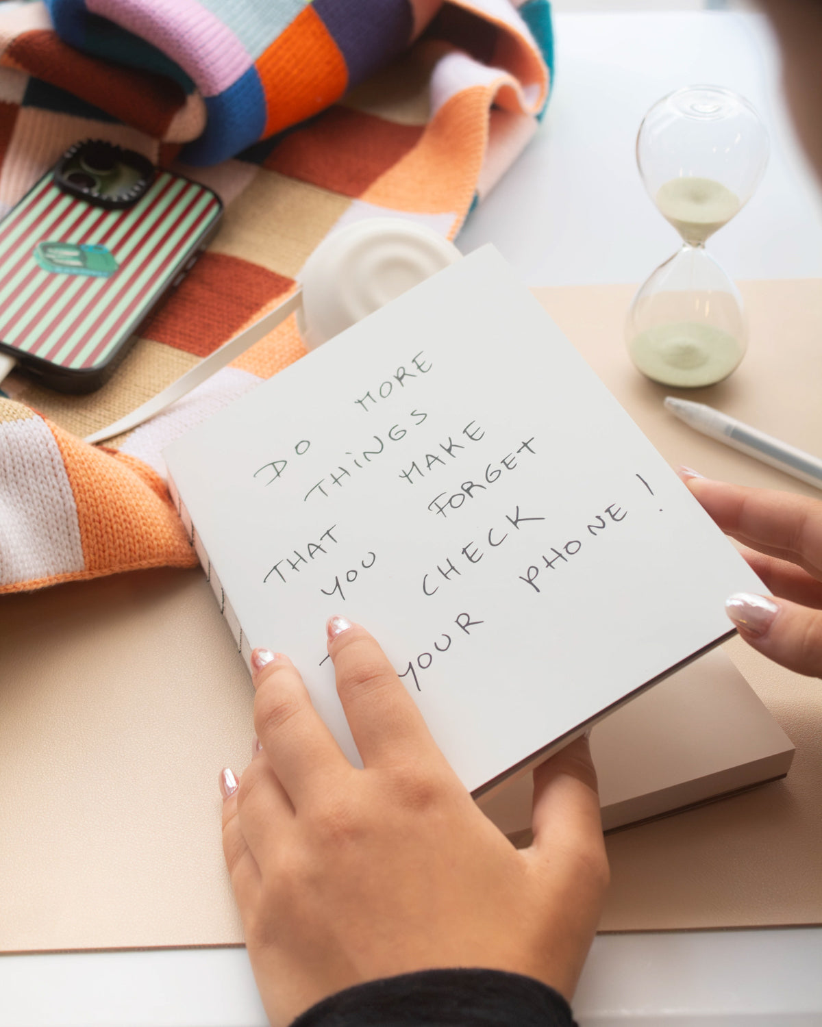 Hand holding the white Poète notebook on a desk with an hourglass and phone