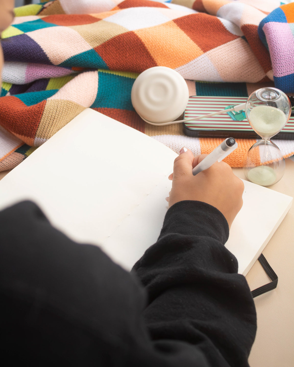 Hand writing in an open lay-flat Poète notebook beside an hourglass and a charger