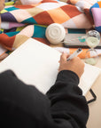 Hand writing in an open lay-flat Poète notebook beside an hourglass and a charger