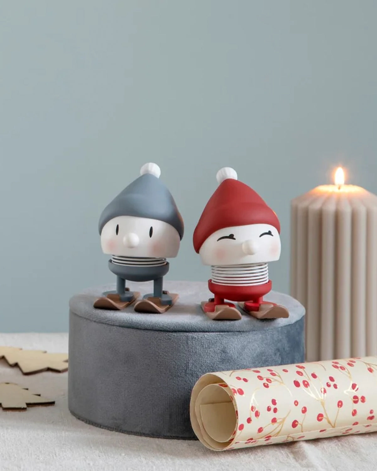 Two Hoptimist skiing figures in blue and red knit caps on a gray stand beside a candle
