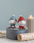 Two Hoptimist skiing figures in blue and red knit caps on a gray stand beside a candle