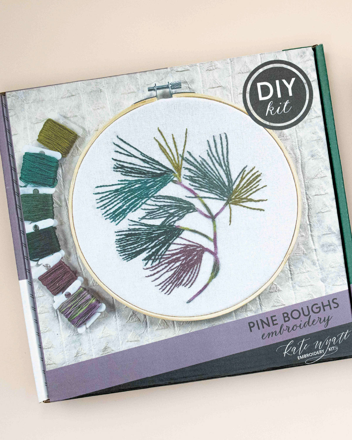 Kate Wyatt Pine Boughs embroidery kit box with a stitched pine branch in a wooden hoop