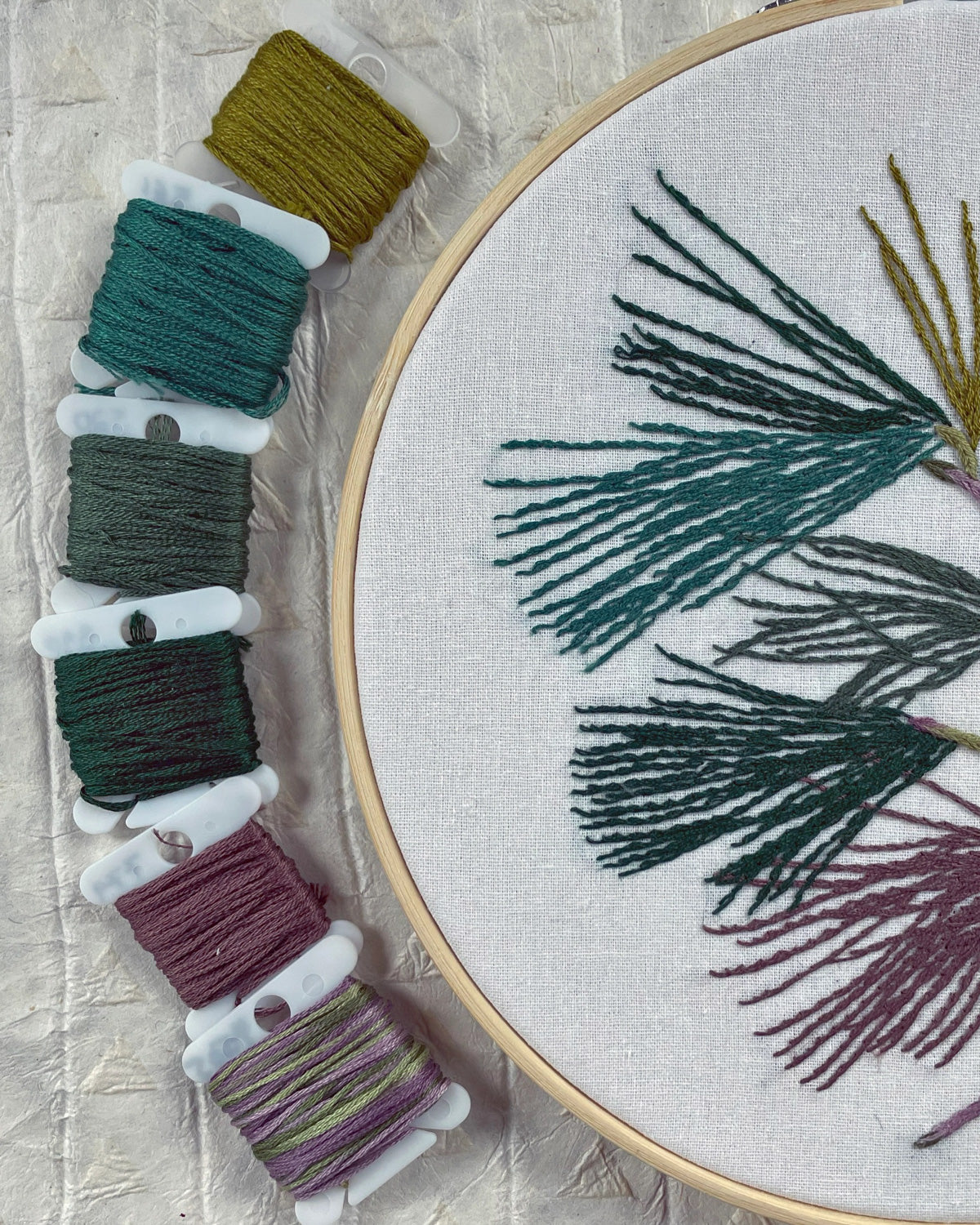 Kate Wyatt pine branch embroidery in progress beside bobbins of green and gray floss