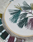 Pine Boughs embroidery hoop next to a row of floss bobbins in greens and plums