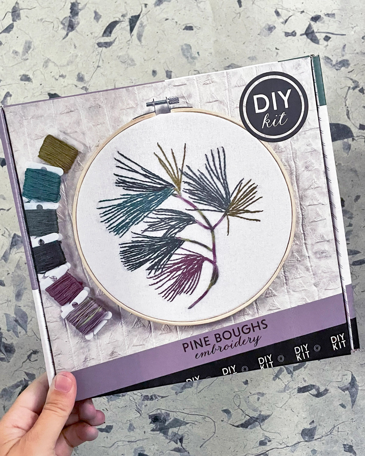 Hand holding the Kate Wyatt Pine Boughs DIY embroidery kit box