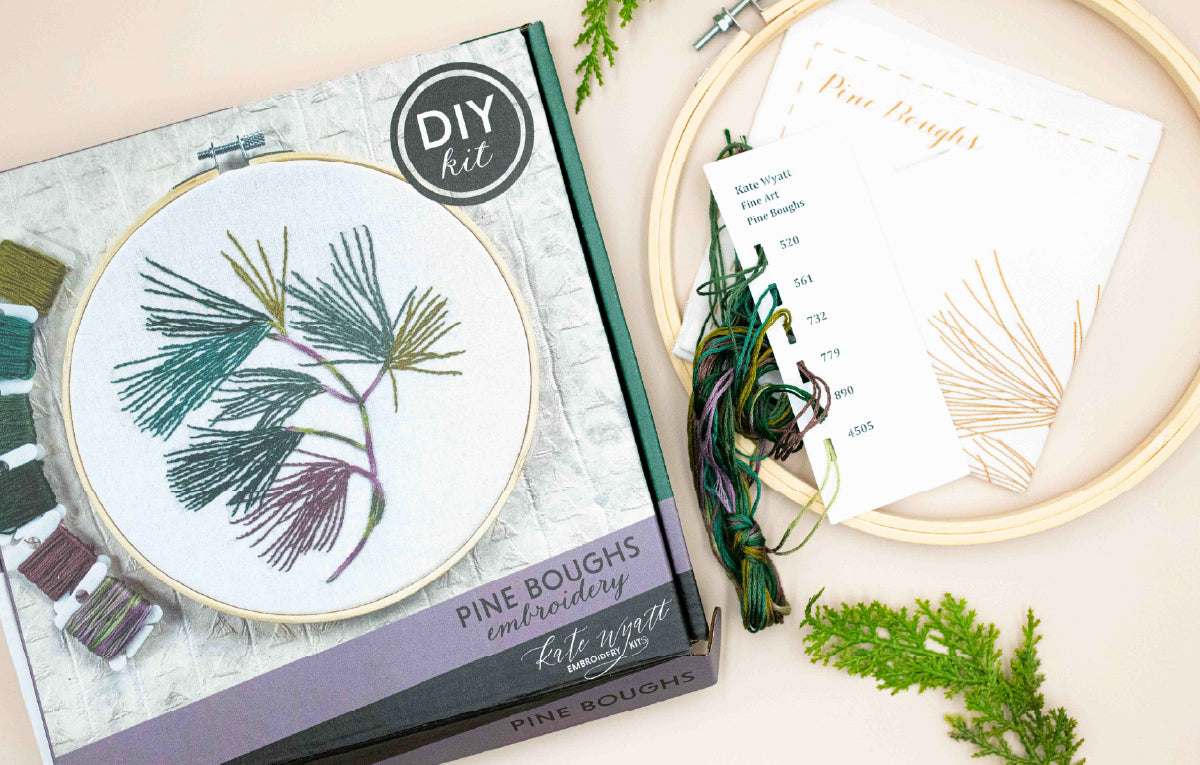 Pine Boughs embroidery kit box beside its stitched hoop and instruction sheet
