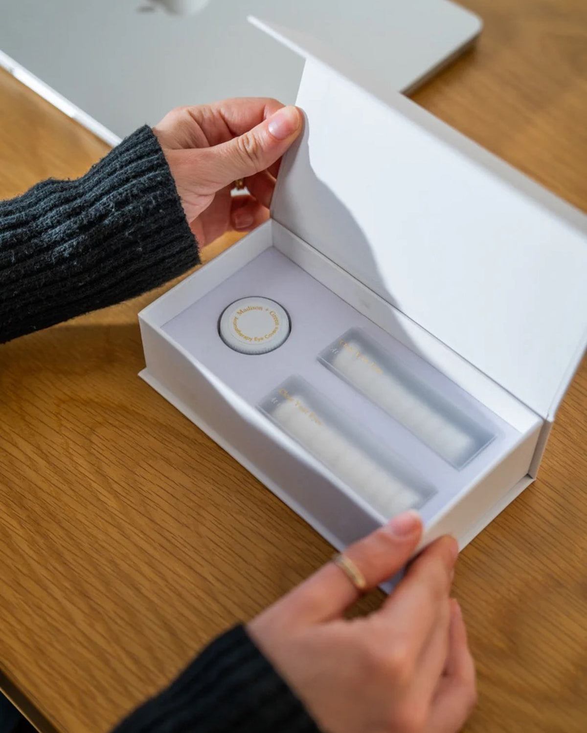 Person opening a gift box of 'Rest Your Eyes' aromatherapy eye towels at a wooden desk.