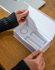 Person opening a gift box of 'Rest Your Eyes' aromatherapy eye towels at a wooden desk.