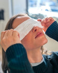 Person wearing a 'Rest Your Eyes' aromatherapy eye towel in an indoor, home setting.
