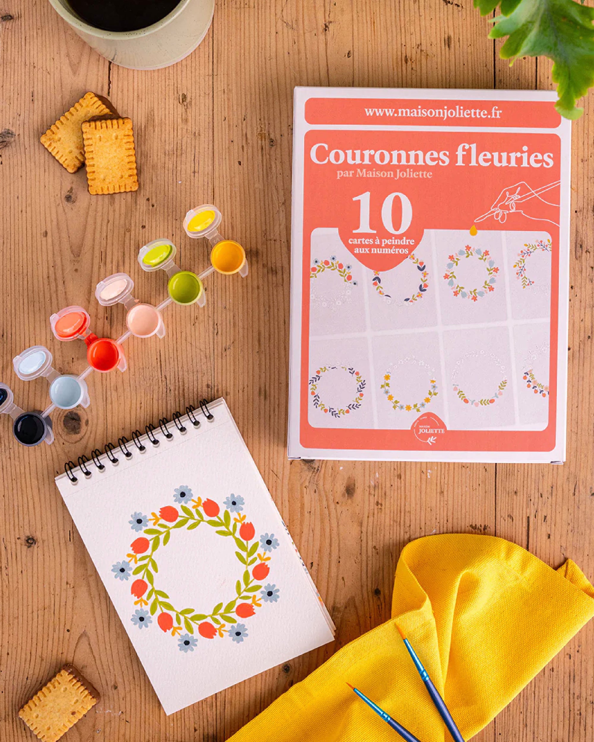 Packaging of 'Couronnes fleuries' by Maison Juliette on a wooden surface with a notebook feutruing a floral wreathe design and art supplies.