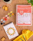 Packaging of 'Couronnes fleuries' by Maison Juliette on a wooden surface with a notebook feutruing a floral wreathe design and art supplies.